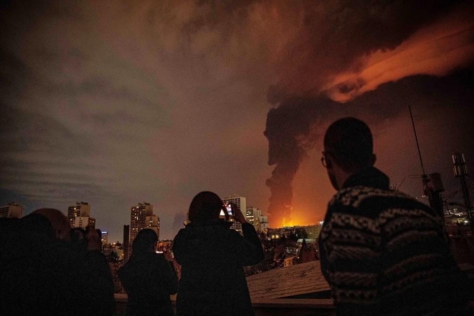Residents watch and take pictures as flames and smoke rise from an oil storage facility struck as attacks hit the city during the U.S.–Israeli military campaign in Tehran, Iran, Saturday, March 7, 2026.